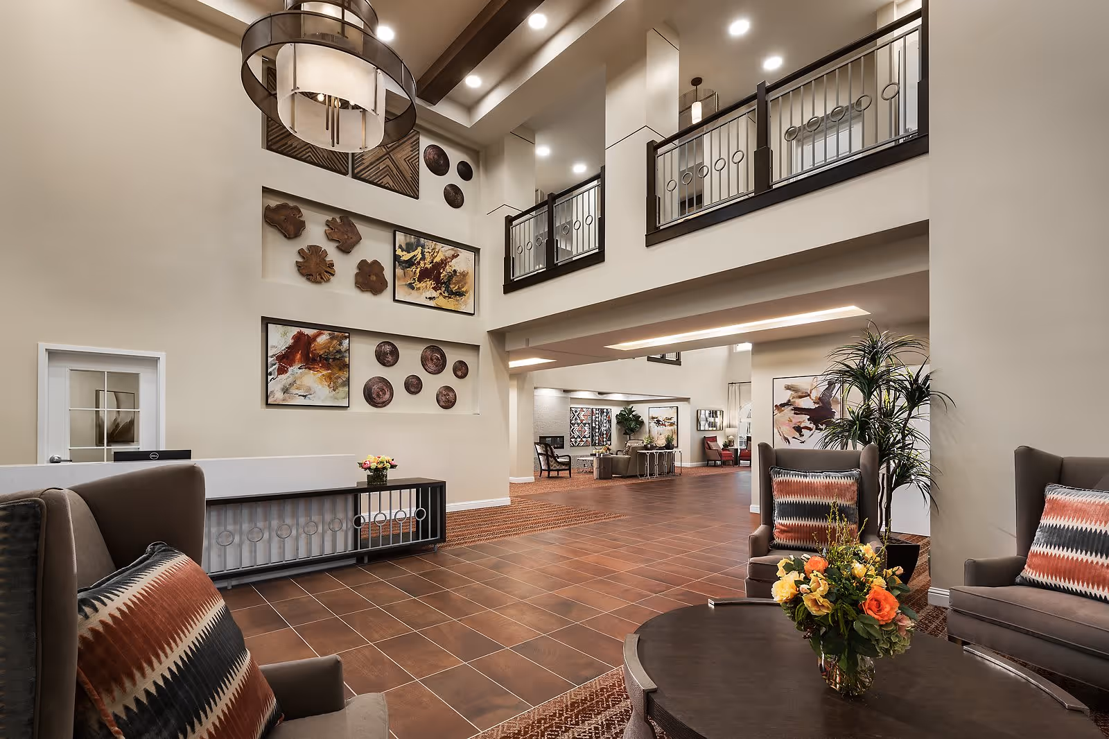 Spacious senior living facility lobby with high ceilings, modern chandelier, decorative wall art, and comfortable seating areas featuring armchairs with patterned pillows and a round table with a floral arrangement.