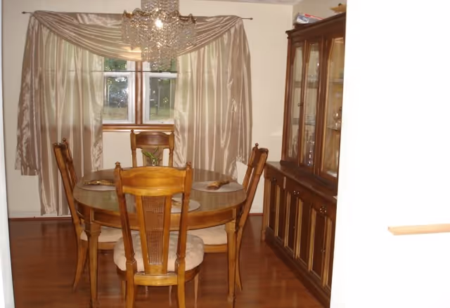 A dining room with a round wooden table surrounded by four wooden chairs with cushioned seats. A chandelier hangs above the table. Behind the table is a window with beige curtains. To the right is a wooden china cabinet with glass doors displaying dishes and glassware.