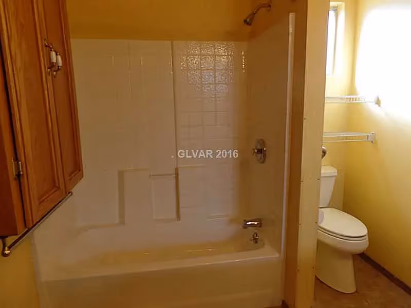 A bathroom with a white bathtub and shower combination, beige tiled walls around the tub, a toilet next to the tub, wooden cabinets on the left wall, and a small window letting in natural light.