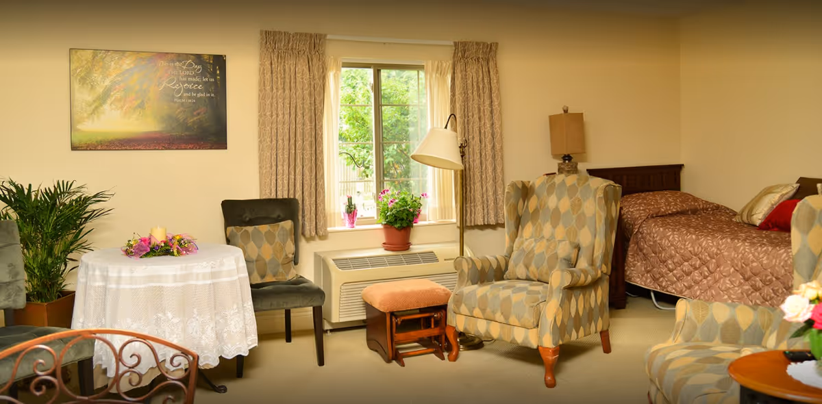 A cozy room with a single bed covered in a brown quilt, two patterned armchairs, a small table with a white lace tablecloth and floral centerpiece, a window with beige curtains, a floor lamp, and a potted plant on the windowsill.