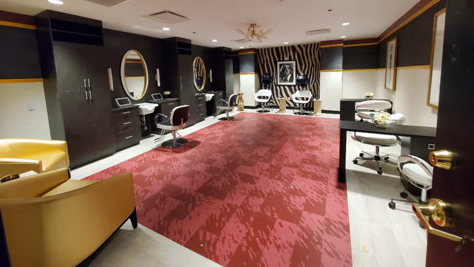 Interior view of a stylish salon room with multiple salon chairs, mirrors, and hair drying stations. The room features dark cabinetry, a red patterned carpet, and a zebra-striped accent wall with a black and white portrait. There are also yellow armchairs and small tables with floral decorations.