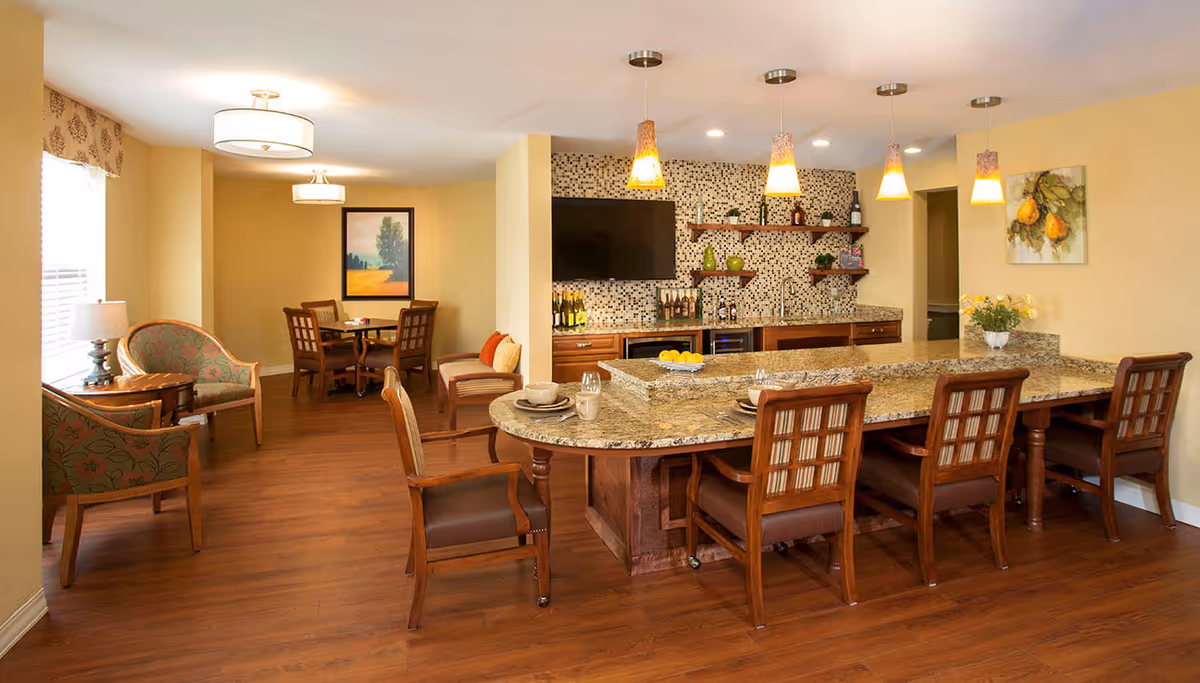 A warm and inviting interior space featuring a granite countertop bar with four wooden chairs, a small dining area with a round table and four chairs, and a cozy sitting area with two upholstered armchairs and a side table with a lamp. The back wall has a mosaic tile backsplash, open wooden shelves with decorative items, a mounted flat-screen TV, and a small sink area. The room has wooden flooring, soft yellow walls, and is well-lit with ceiling lights and pendant lamps.