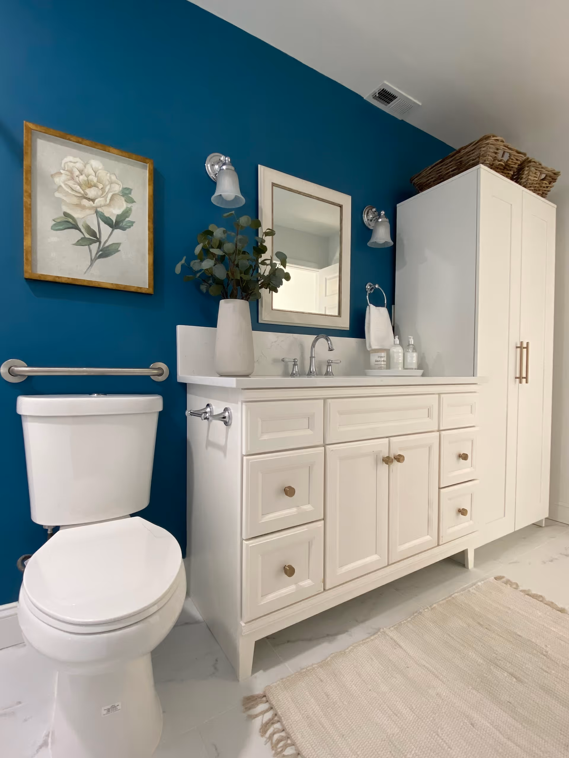A clean and modern bathroom featuring a white toilet, a white vanity with multiple drawers and cabinets, a rectangular mirror above the sink, two wall-mounted light fixtures, a vase with green foliage on the vanity, a towel ring with a white hand towel, a framed floral artwork on a teal accent wall, a tall white cabinet with wicker baskets on top, and a light-colored rug on the floor.