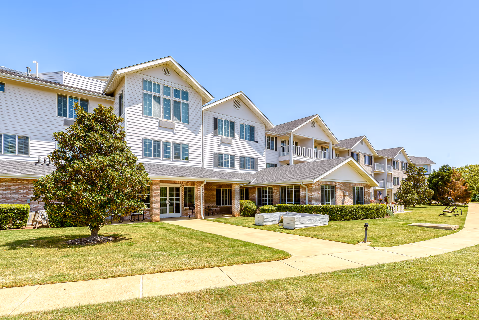 Exterior view of a multi-story senior living facility building with white siding and brick accents, surrounded by a well-maintained lawn, trees, and paved walkways under a clear blue sky.