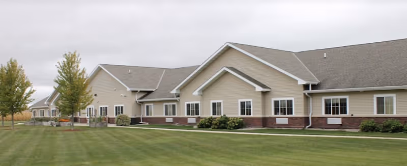 Exterior view of a single-story beige building with multiple windows, a sloped roof, and a well-maintained green lawn with a few trees in front under a cloudy sky.
