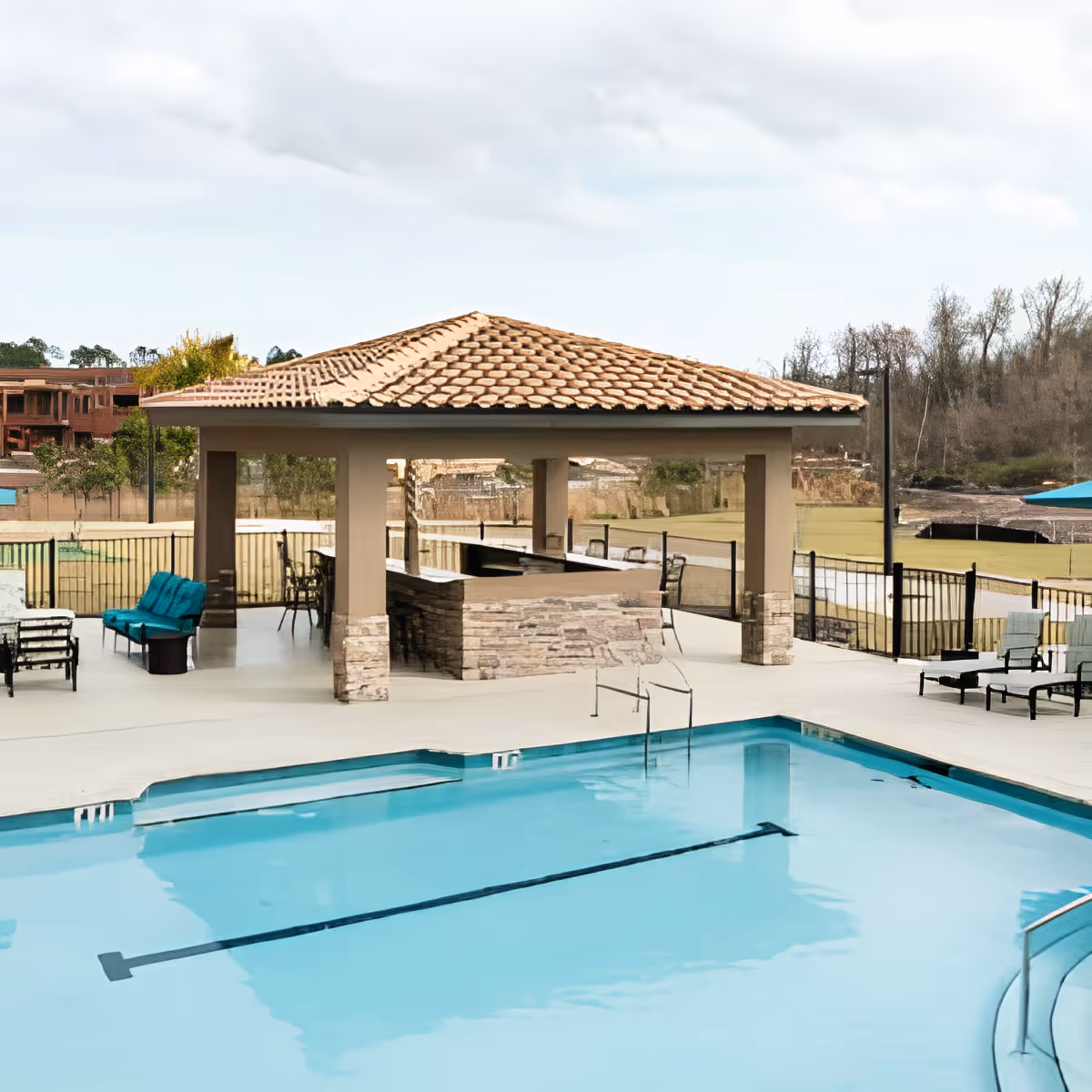 Outdoor swimming pool area with clear blue water, surrounded by a concrete deck with lounge chairs and a covered pavilion with a tiled roof and stone pillars. Trees and a grassy area are visible in the background under a cloudy sky.