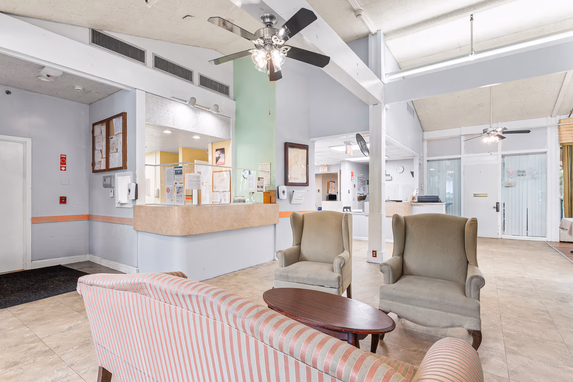 A bright and spacious reception area in a senior living facility with a striped sofa, two beige armchairs, a wooden coffee table, ceiling fans, and a reception desk with protective glass panels. The room has tiled floors, light-colored walls, and large windows allowing natural light.