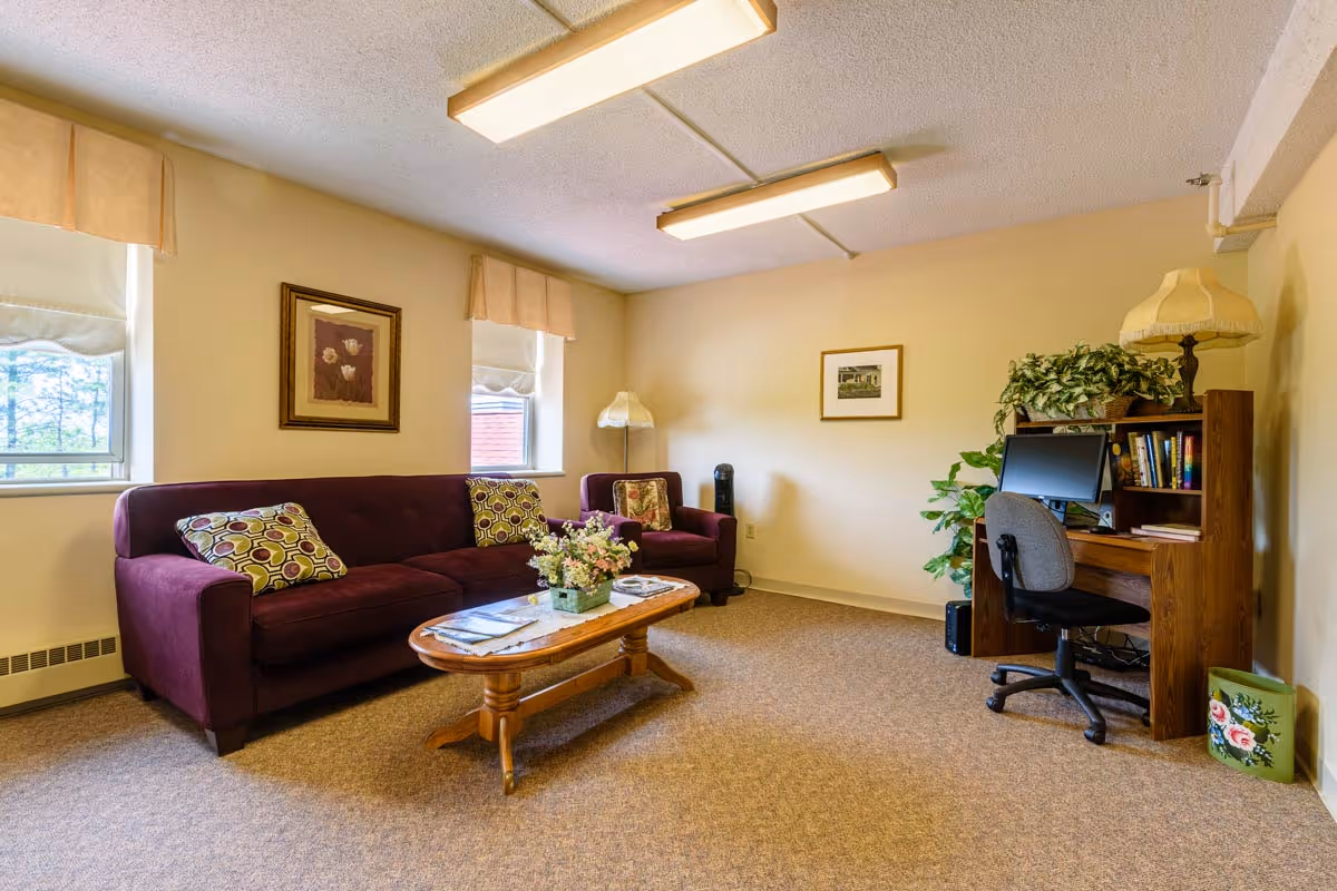 A cozy living room area with a purple sofa and matching armchair, a wooden coffee table with a flower arrangement and magazines, two windows with beige curtains, framed artwork on the walls, and a wooden desk with a computer, chair, lamp, and plants.