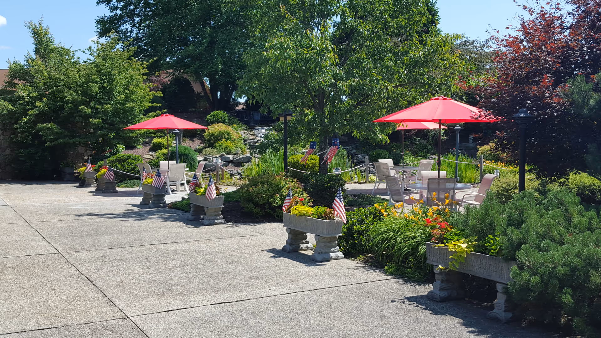 Outdoor patio area with several round tables and chairs under red umbrellas surrounded by lush greenery and flower planters decorated with small American flags. Trees and bushes provide shade and a peaceful garden atmosphere.