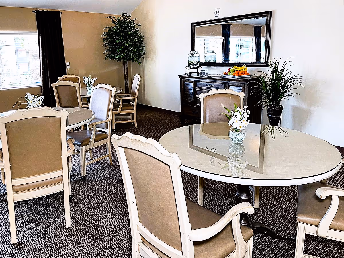 Small dining area with round glass-topped tables and upholstered chairs, a sideboard with a mirror, and decorative plants.