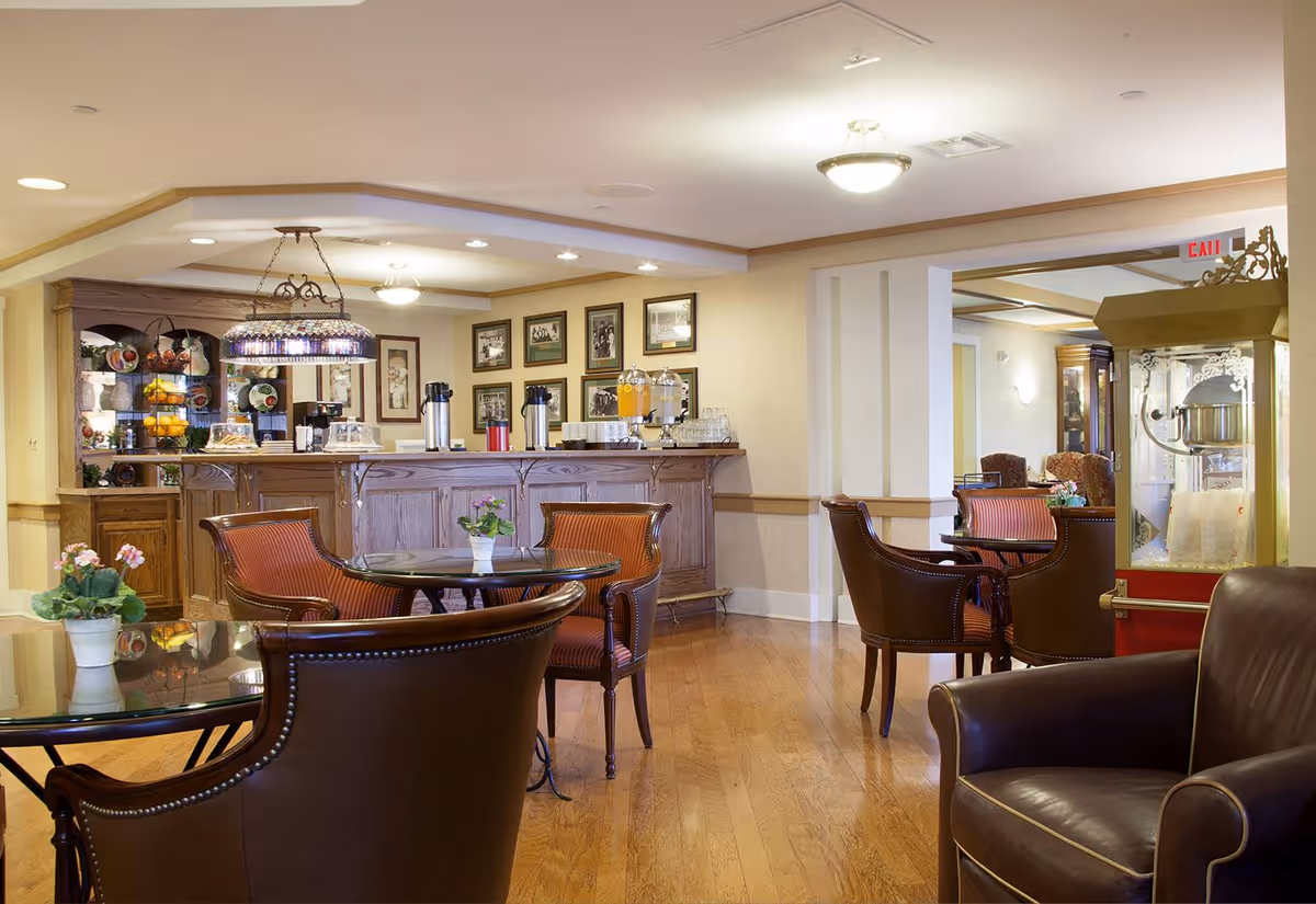 Well-lit senior living common area with round glass tables, leather chairs, a wooden service counter with beverage dispensers and a popcorn machine.