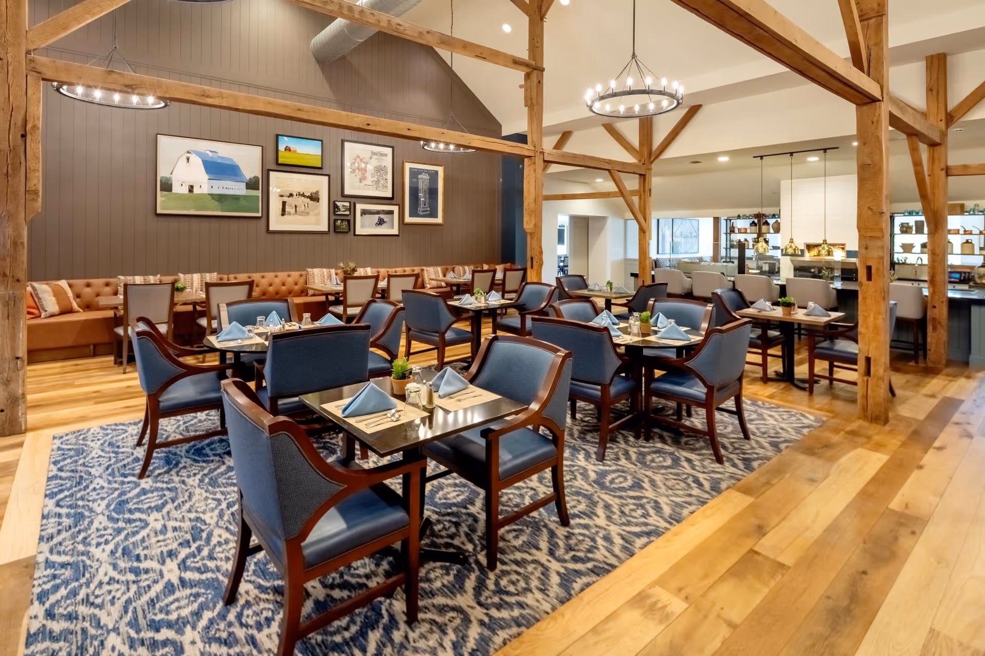 A spacious dining room with wooden beams and high ceilings. The room features multiple tables set with blue napkins, silverware, and small potted plants. Blue upholstered chairs surround the tables, and a patterned blue and white rug covers part of the wooden floor. The back wall has a brown panel with framed artwork, and there is a long cushioned bench along the wall. Pendant lights hang over a counter area in the background.