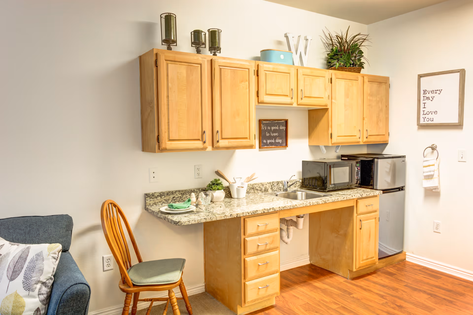 Small kitchenette with wooden cabinets, granite countertop, sink, microwave and mini-fridge next to a chair and partial sofa.