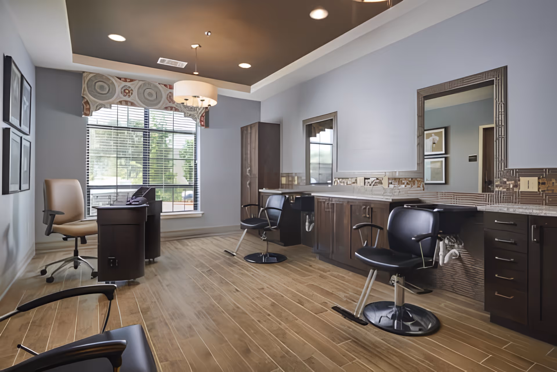 Interior view of a salon area in an assisted living facility featuring wooden flooring, two black salon chairs in front of a long counter with mirrors, a desk with a beige office chair near a large window with blinds and decorative valance, and framed artwork on the wall.