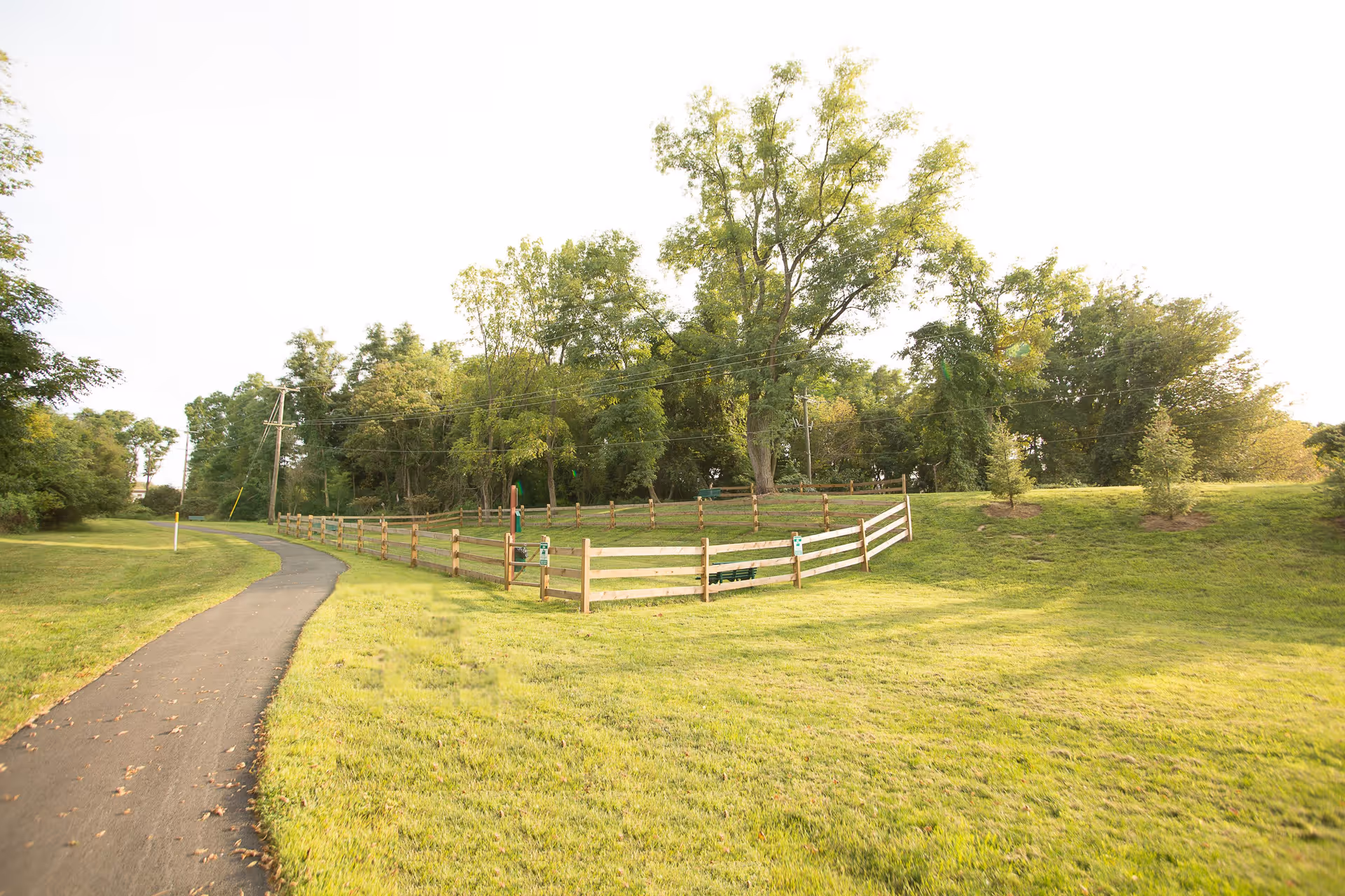 A paved walking path curves past a grassy field with a wooden fence and trees in the background.