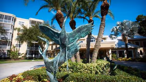 Exterior view of Royal Palms facility with palm trees and a bird sculpture.