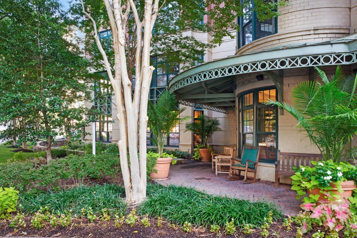 Outdoor patio area of a senior living facility with potted plants, wooden benches, and rocking chairs under a curved metal awning attached to a brick building. The surrounding area has green shrubs, trees, and landscaped garden beds.