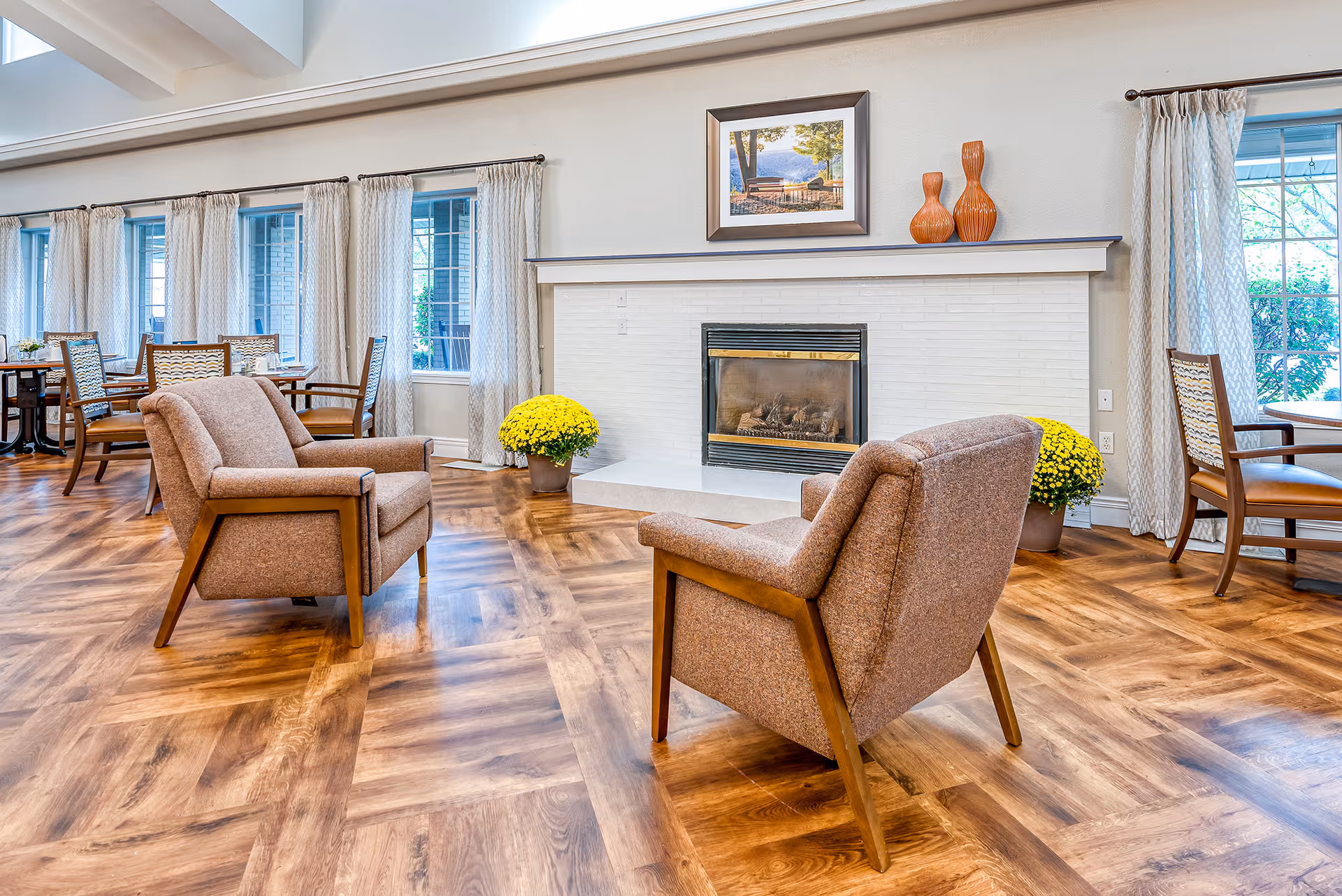 Communal senior living lounge with two upholstered armchairs facing a fireplace, wood-patterned floor, dining tables, and large windows.