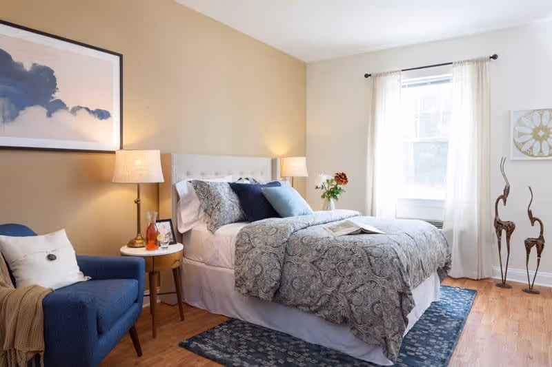 A cozy bedroom with a neatly made bed featuring patterned bedding and multiple pillows. There is a blue armchair with a white pillow and a beige throw blanket next to a small round wooden side table with a lamp, a glass bottle, and a framed photo. The room has wooden flooring, a blue patterned rug, a window with sheer white curtains, and decorative sculptures and artwork on the walls.