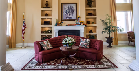 A cozy living room area with a curved maroon sofa adorned with floral cushions, centered on a patterned rug. In front of the sofa is a round wooden coffee table with a floral arrangement. Behind the sofa is a white fireplace with built-in shelves on either side holding decorative items and books. Above the fireplace hangs a framed floral painting. The room has tall windows with beige curtains, a potted plant on the right, and an American flag on the left.