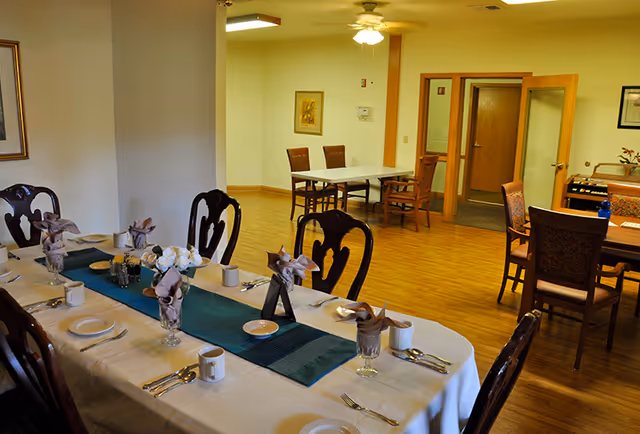 Dining room with a long set table laid with napkins, plates and cups, with additional tables and chairs on hardwood floors.