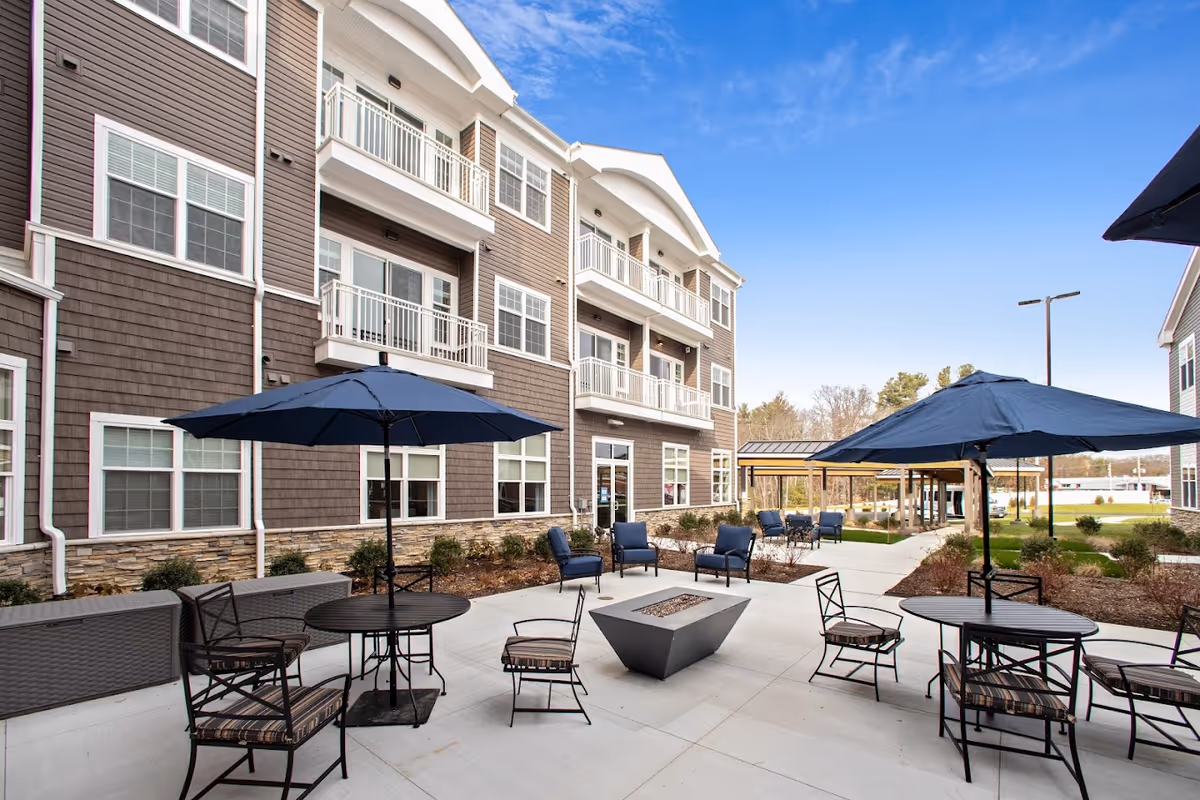Outdoor patio area at Artisan at Hudson with multiple round tables and chairs, each table shaded by a large navy blue umbrella. The patio is adjacent to a multi-story building with balconies and large windows. There is a modern fire pit in the center of the patio and a covered seating area in the background. The sky is clear and blue.