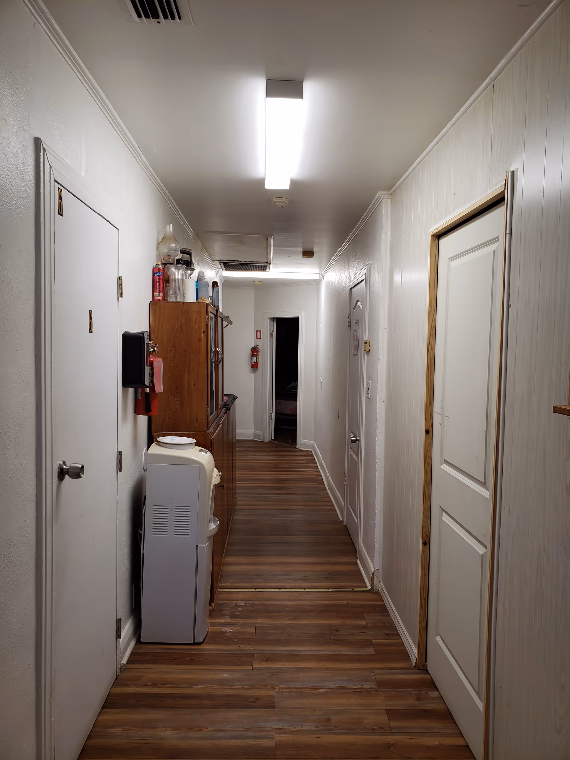 Narrow indoor hallway with wood flooring, doors on both sides, a water cooler and cabinets under fluorescent ceiling lights.