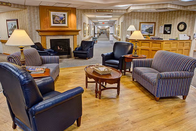 A cozy seating area in a senior living facility with two striped sofas, two dark leather armchairs, a wooden coffee table with a basket, and two table lamps. In the background, there is a fireplace with a painting above it, a reception desk, and a long hallway with framed pictures on the walls.