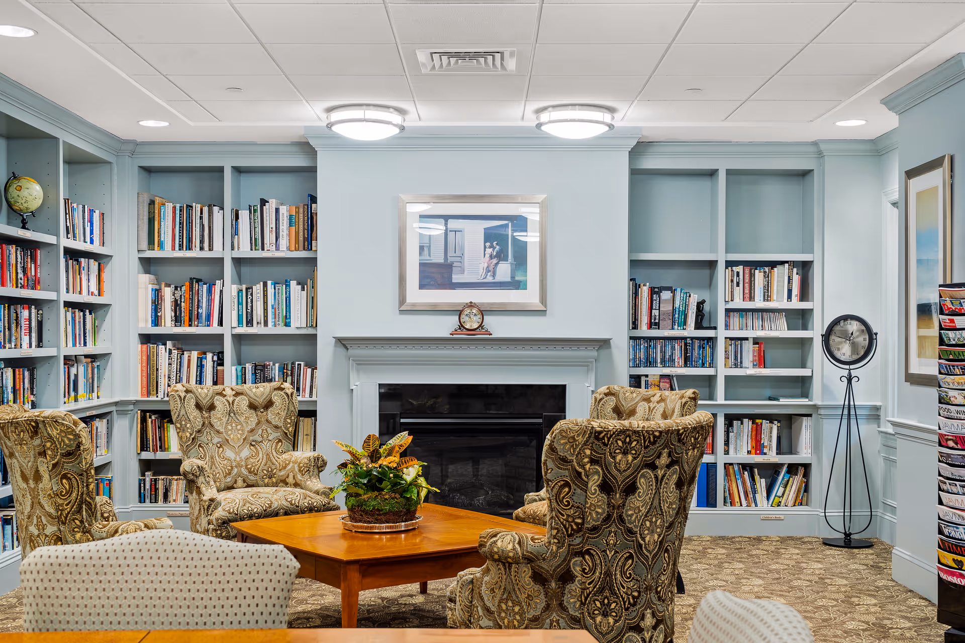 A cozy library room with built-in light blue bookshelves filled with books surrounding a fireplace. Four patterned armchairs are arranged around a wooden coffee table with a plant centerpiece. A framed picture hangs above the fireplace, and a standing clock is positioned near the right bookshelf. The room has a carpeted floor and a white ceiling with recessed lighting.
