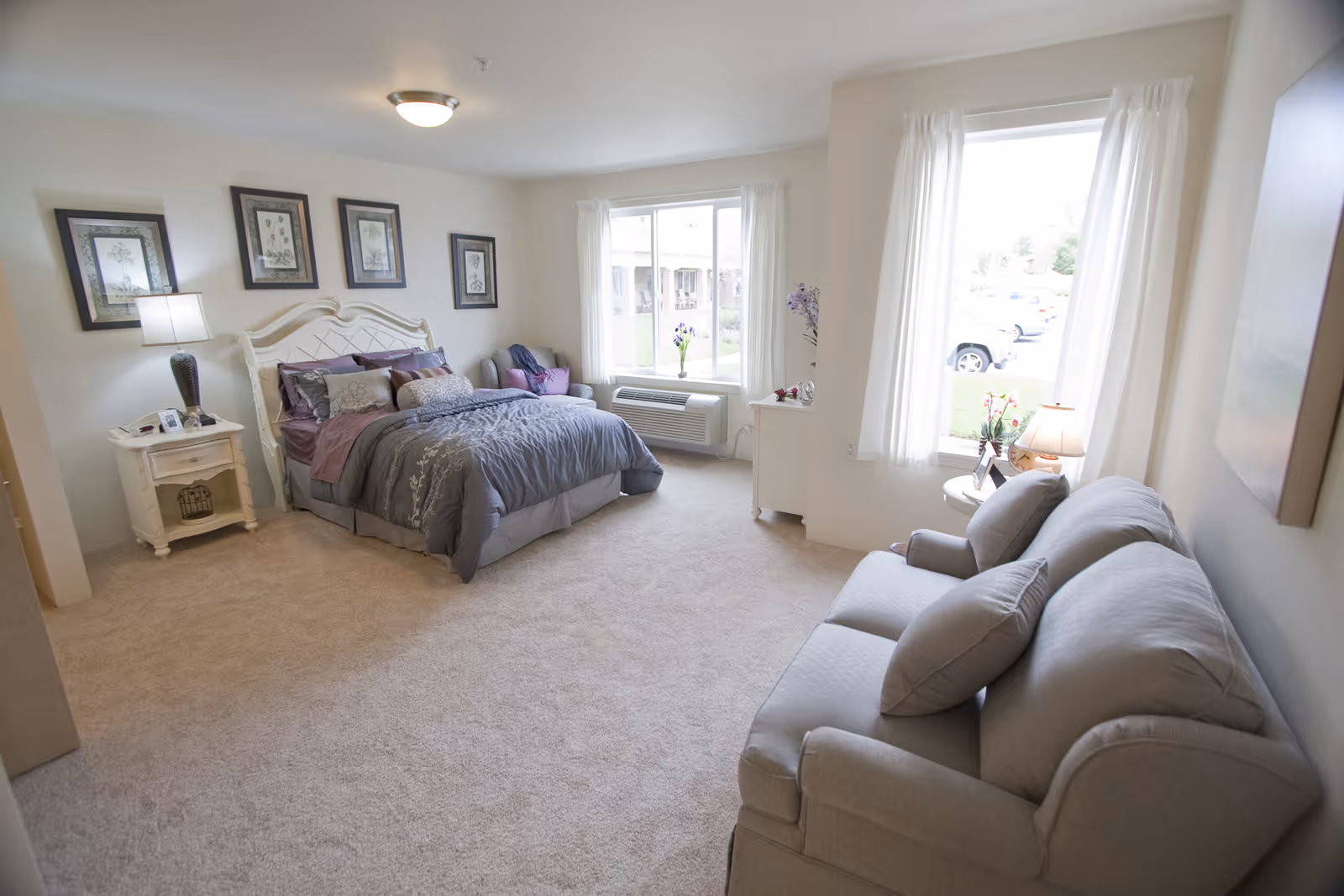 A bright and spacious senior living bedroom with a large bed adorned with gray and purple bedding, a nightstand with a lamp and picture frame, a small armchair near the window, a dresser with flowers, and a comfortable gray sofa. The room has beige carpet, white walls, and large windows with white curtains letting in natural light.