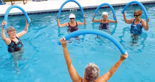 A group of elderly women participating in a water exercise class in an outdoor swimming pool, each holding a blue pool noodle above their heads.