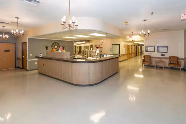 Reception area with a large curved nurses' station, chandeliers, hallway and seating in a senior living facility.