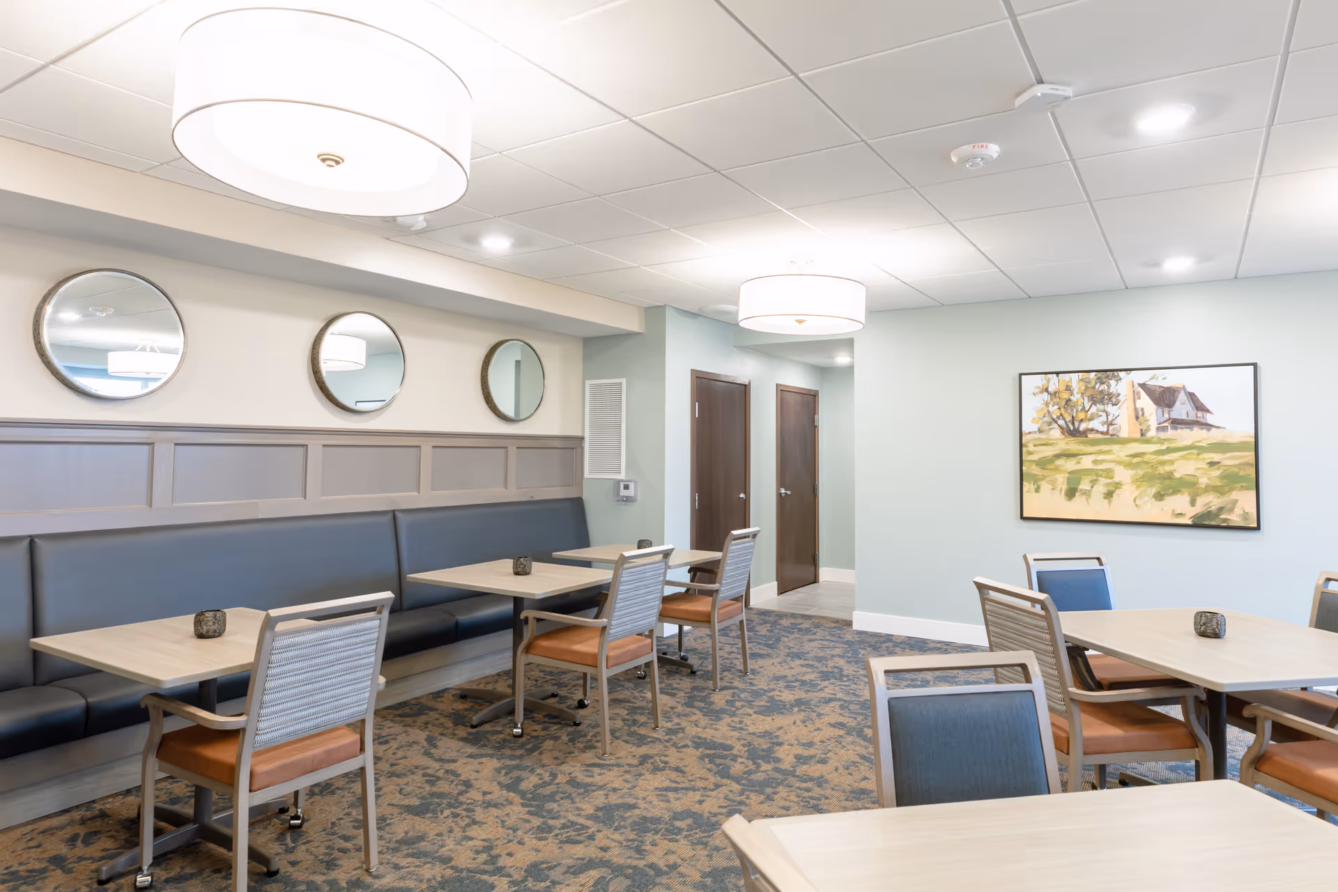 A bright dining area with light wood tables and chairs featuring blue and brown cushions. There is a long black cushioned bench along one wall with three round mirrors above it. The walls are painted light blue and beige, with a framed painting of a house and trees on one wall. The ceiling has recessed lighting and two large round pendant lights. The floor is covered with a patterned carpet.