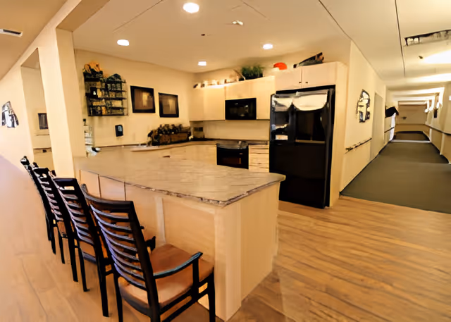 Communal kitchen with a long marble island, row of barstools, refrigerator and cabinets opening onto a hallway.