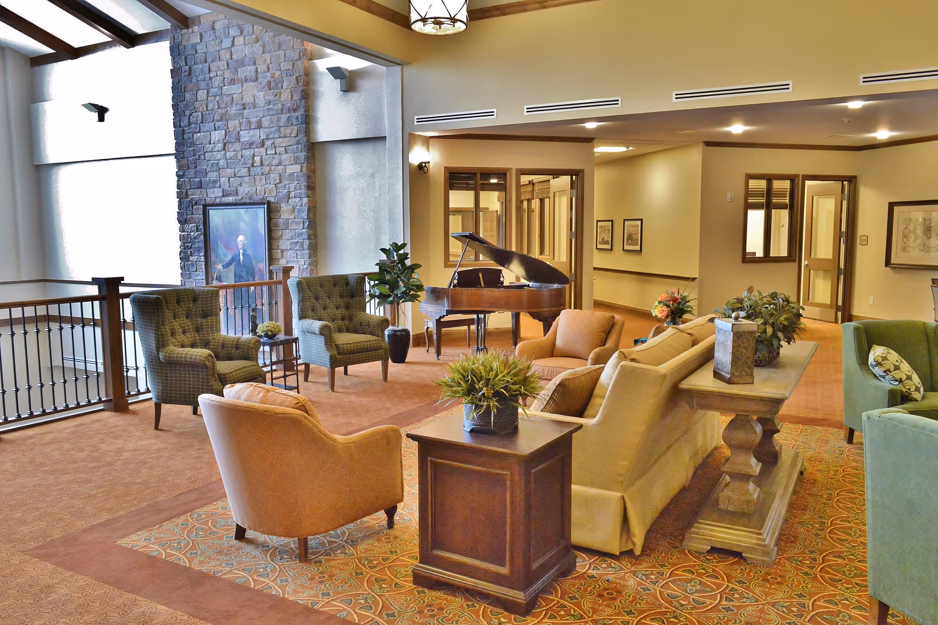 A spacious and well-lit common area in a senior living facility featuring comfortable armchairs and sofas arranged around a coffee table with potted plants. A grand piano is positioned near a stone accent wall with a framed portrait. The room has warm lighting, carpeted floors, and decorative elements including framed artwork and plants.