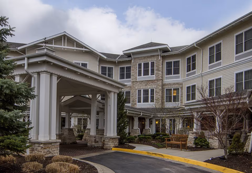 Exterior view of a multi-story senior living facility with beige siding and stone accents. The building features many windows and a covered entrance supported by white columns with stone bases. There are landscaped areas with shrubs, trees, and a wooden bench near the entrance. The sky is partly cloudy.