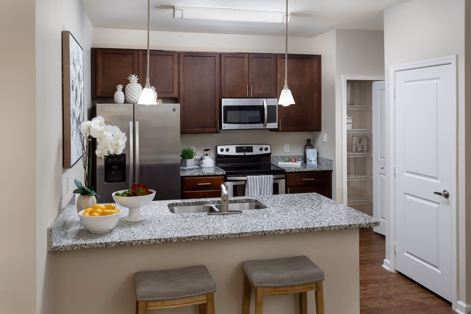 Modern kitchen with granite countertops, dark wooden cabinets, stainless steel refrigerator, microwave, and stove. Two pendant lights hang over the counter with a double sink and two stools. Decorative items include white ceramic pineapples, a bowl of lemons, a bowl of green apples, and a white orchid plant. There is an open pantry door and a closed white door on the right side.