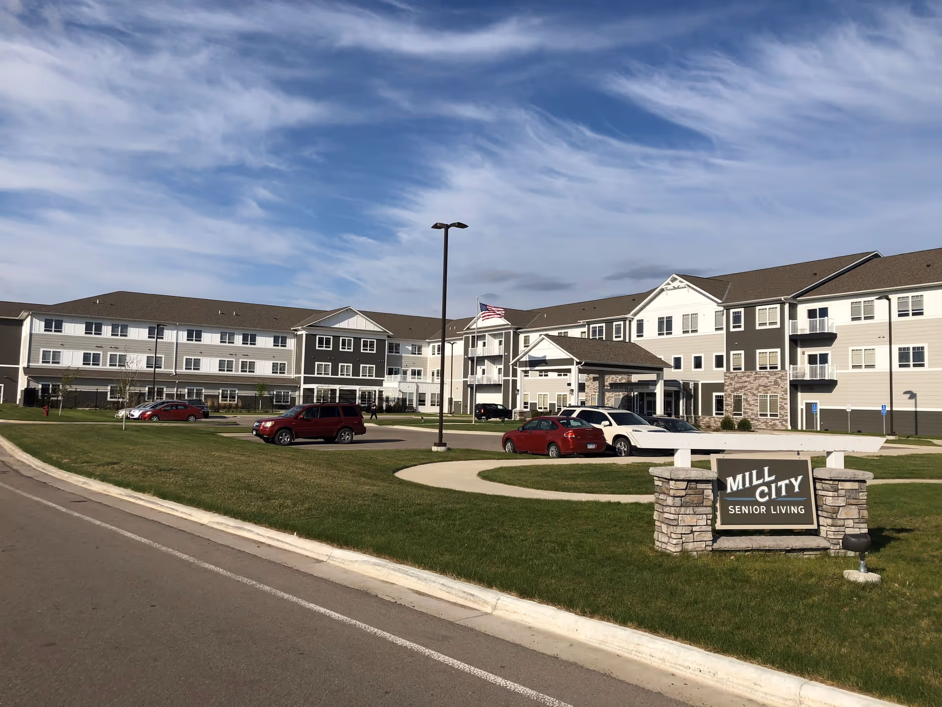 Front exterior of the Mill City Senior Living building with a lawn, sign, parked cars, and an American flag.
