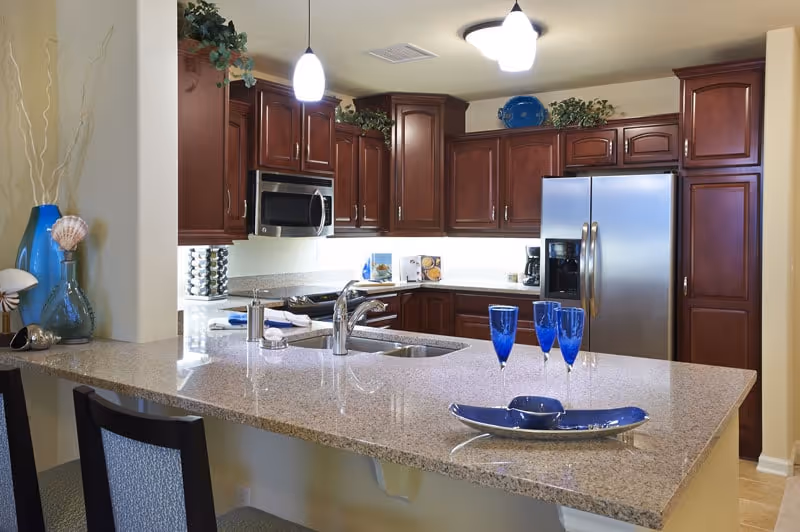 Modern kitchen with a granite island and sink, stainless steel refrigerator and microwave, dark wood cabinets, and blue glassware on the counter.
