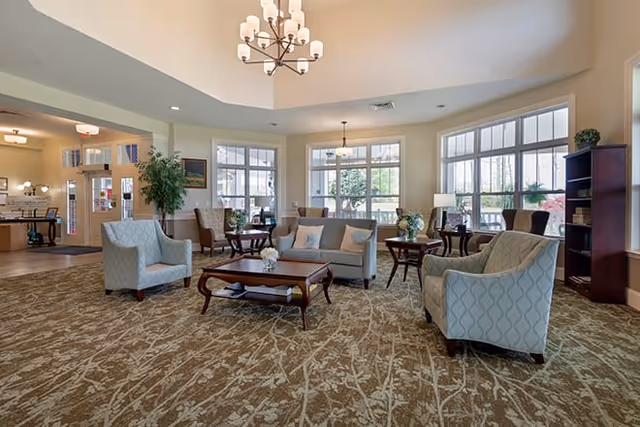 Bright communal living room with sofas and armchairs arranged around coffee tables under a chandelier by large windows.