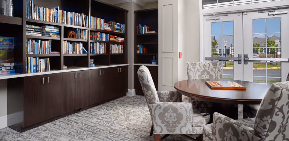 A cozy reading and game area in a senior living facility featuring a large dark wood bookshelf filled with books and board games. In front of the bookshelf is a round wooden table with a checkers game set up, surrounded by four upholstered chairs with a gray and white damask pattern. Double glass doors in the background provide a view of the outside parking area and buildings.