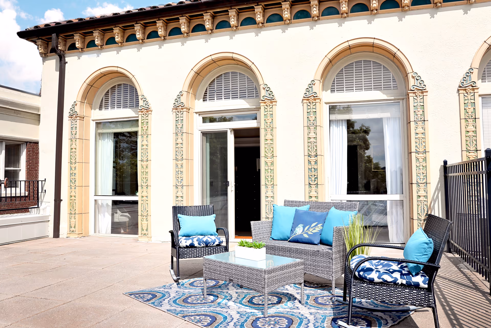 Outdoor patio with wicker seating, blue cushions, a glass-topped coffee table, and arched windows on the building behind.