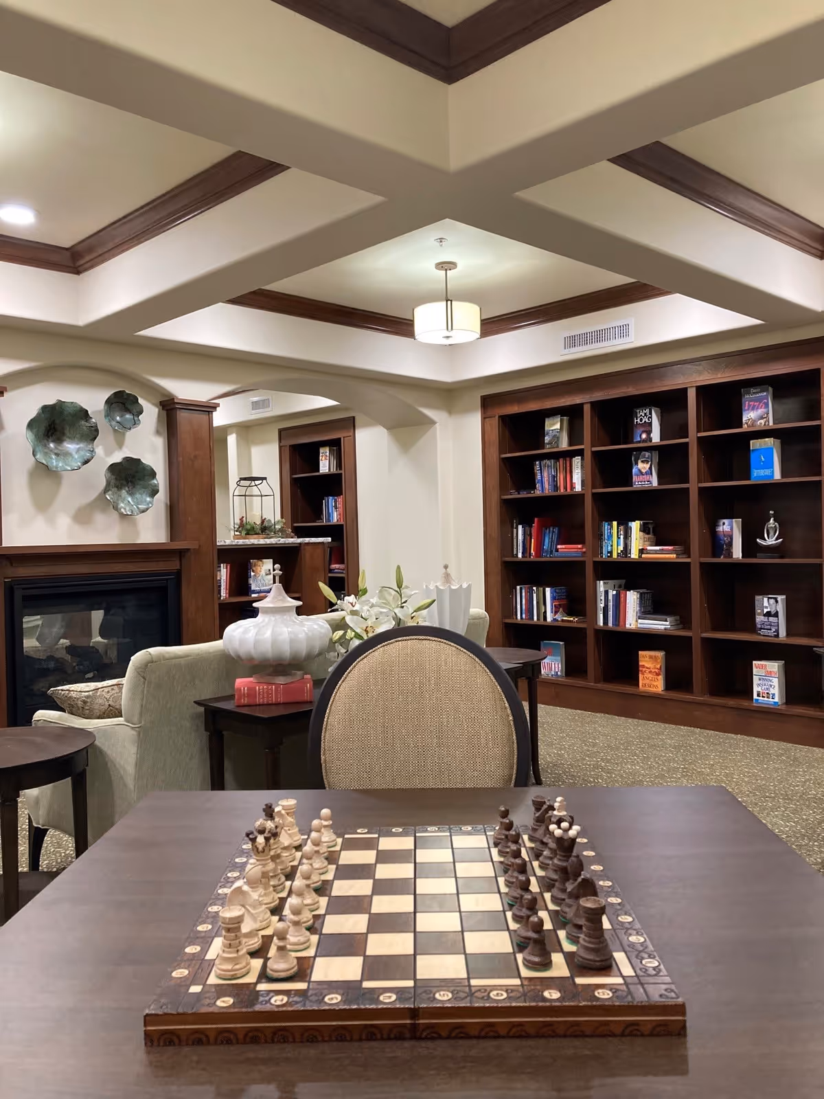 A cozy interior room featuring a chessboard set up on a wooden table in the foreground. Behind the table is a cushioned chair, a small side table with a white decorative vase and flowers, a fireplace with artistic wall decor above it, and built-in wooden bookshelves filled with books. The ceiling has a coffered design with recessed lighting and a central hanging light fixture.