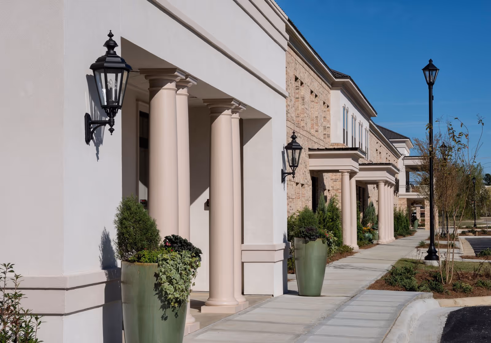 Exterior view of The Blake at Flowood building showing a sidewalk lined with large planters containing greenery, beige columns supporting the building's overhang, brick and stucco walls, and black lantern-style wall lights under a clear blue sky.