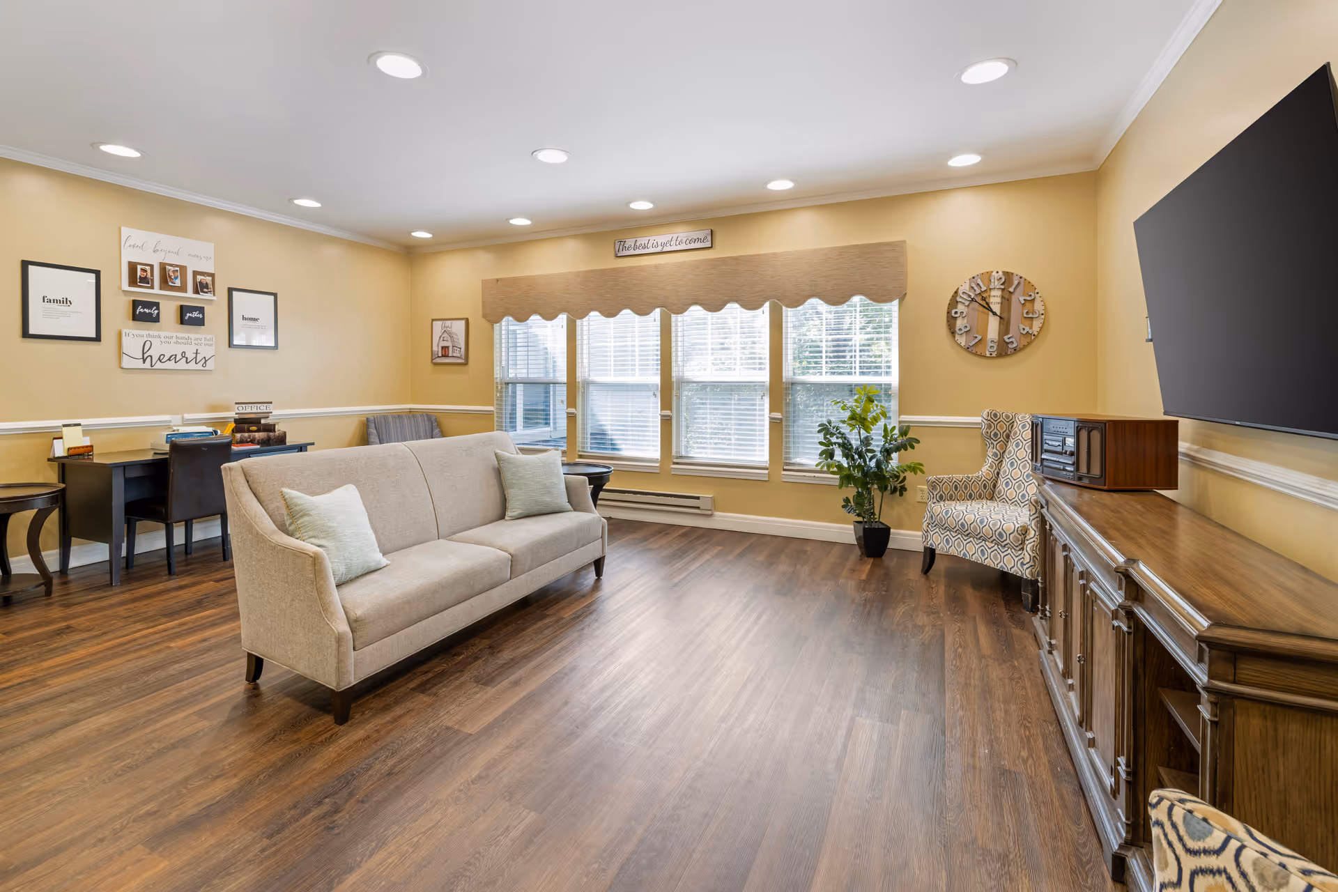 Bright communal living room with a sofa, armchairs, a large TV, wooden floors, and windows letting in natural light.