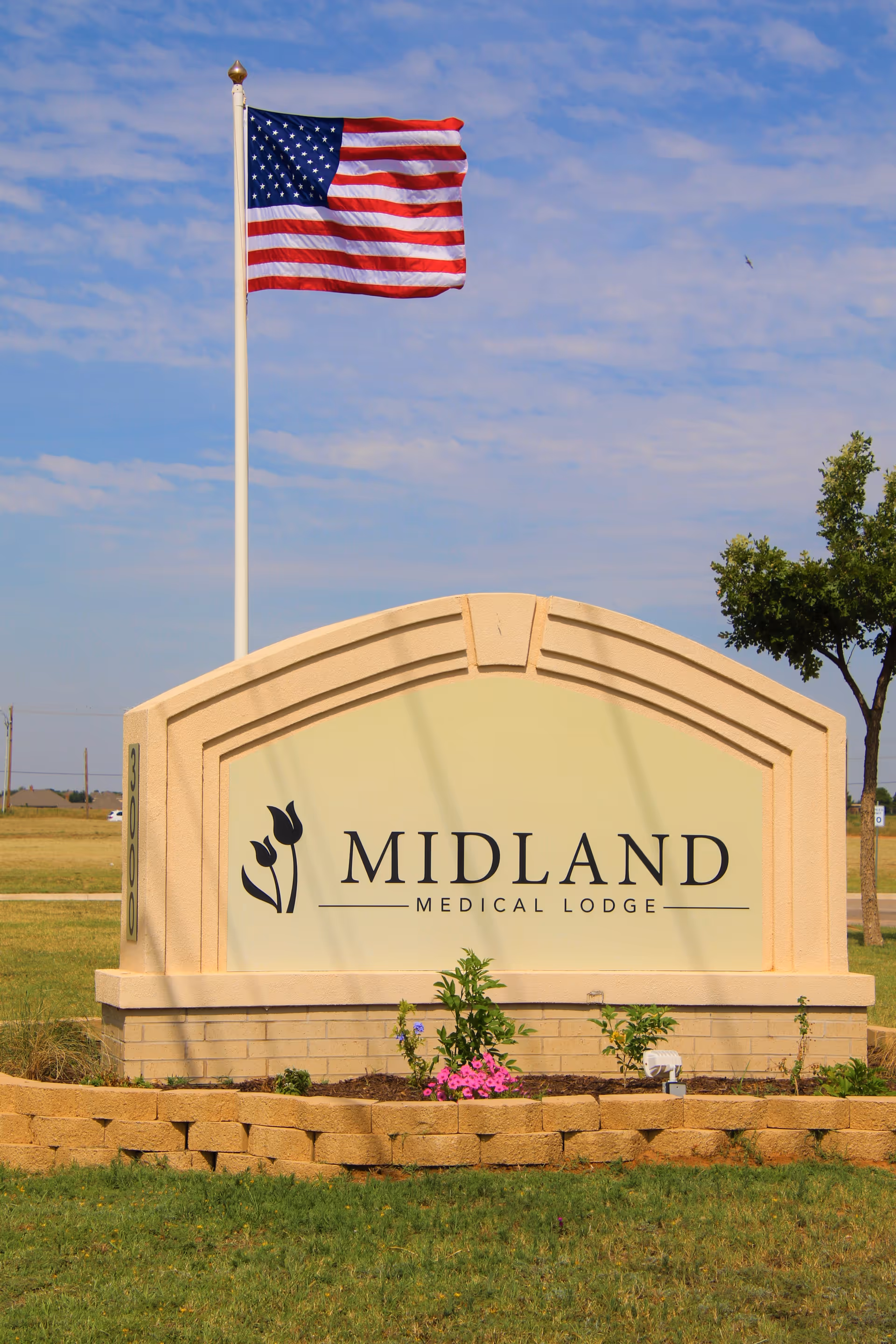Outdoor view of a beige stone sign for Midland Medical Lodge with a flower bed in front and an American flag on a flagpole behind it under a partly cloudy sky.