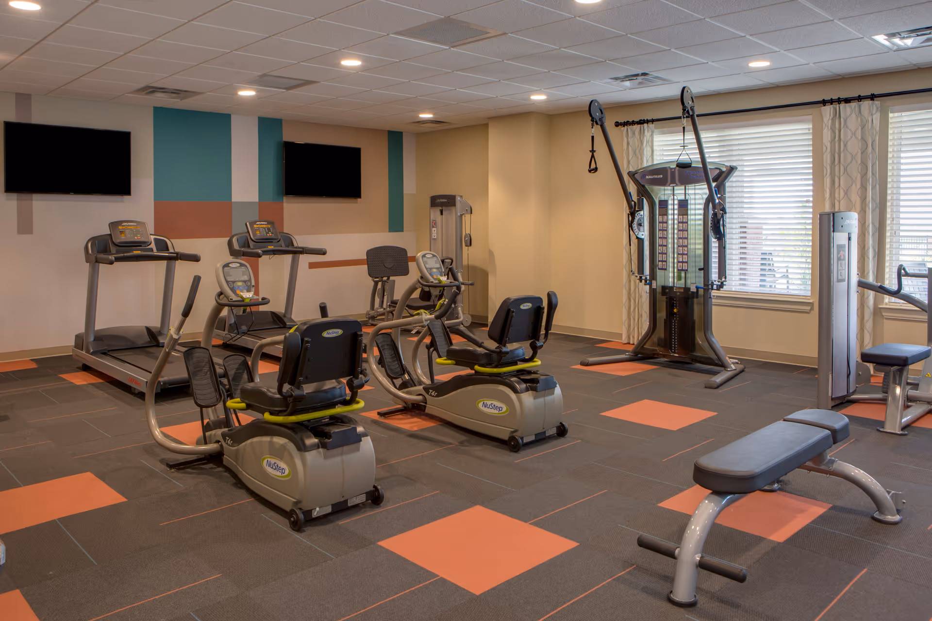 A fitness room with exercise equipment including two treadmills, two recumbent bikes, a multi-functional weight machine, and a bench. The room has a patterned carpet with orange and gray squares, two wall-mounted TVs, and large windows with curtains allowing natural light.