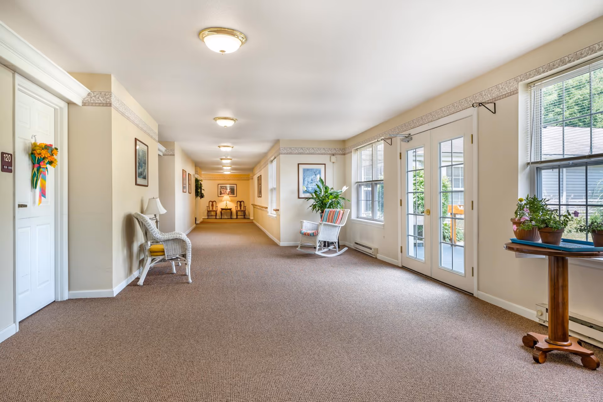 Bright carpeted common hallway with wicker chairs, potted plants, and French doors leading outside.