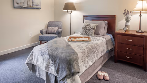 A cozy bedroom with a single bed featuring a floral patterned comforter, a gray throw blanket, and decorative pillows. A wooden nightstand with a lamp, clock, and vase with flowers is beside the bed. A cushioned armchair with a pillow is in the corner, and a pair of pink slippers are placed on the carpeted floor near the bed.