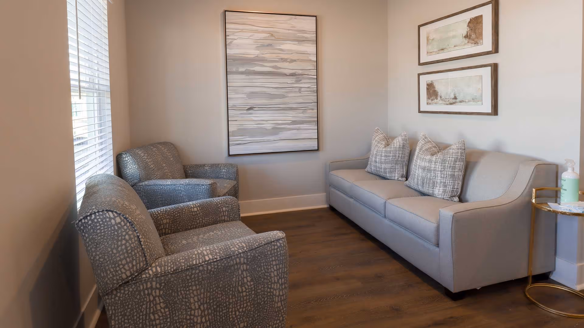 A cozy living room area with a gray sofa adorned with three patterned pillows, two matching armchairs with a textured design, wooden flooring, and light-colored walls decorated with three framed abstract artworks. A small round side table with a bottle of hand sanitizer is placed next to the sofa.